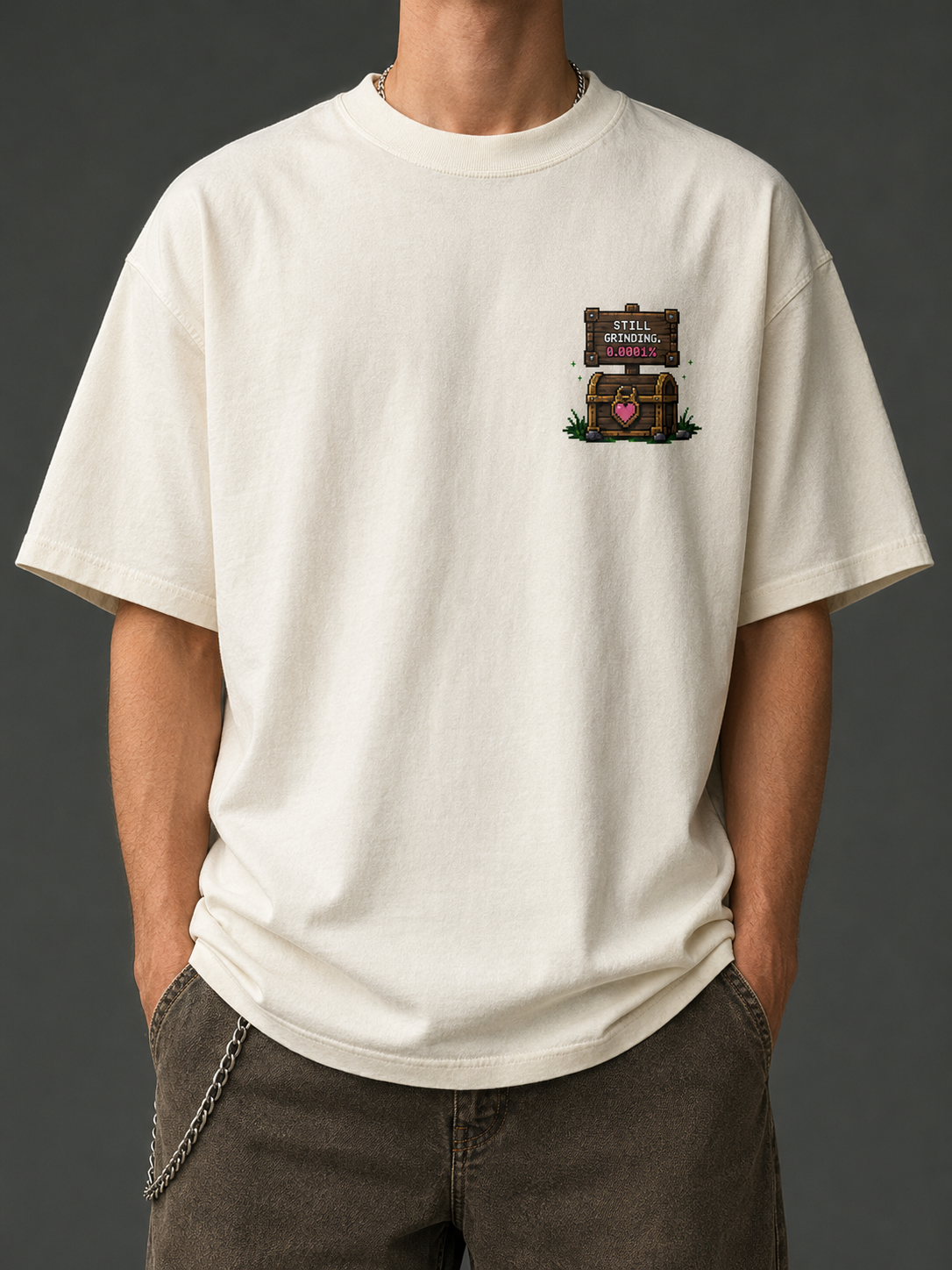 Man wearing the Ruxel Love Drop Rate Cream tee, front view - small pixel Still Grinding sign and heart treasure chest on the left chest