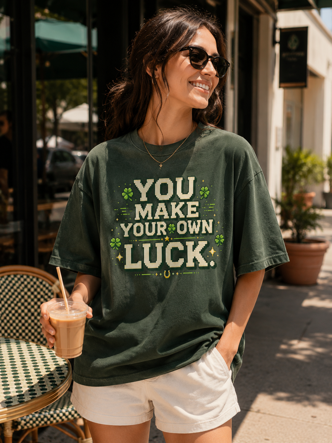 Woman at a cafe holding an iced coffee, wearing the Ruxel You Make Your Own Luck Clover Green pixel-art tee, front view