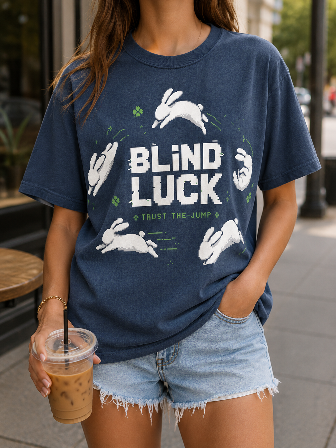 Woman on a city street holding an iced coffee, wearing the Ruxel Blind Luck Midnight Navy pixel-art tee with the leaping rabbit front print