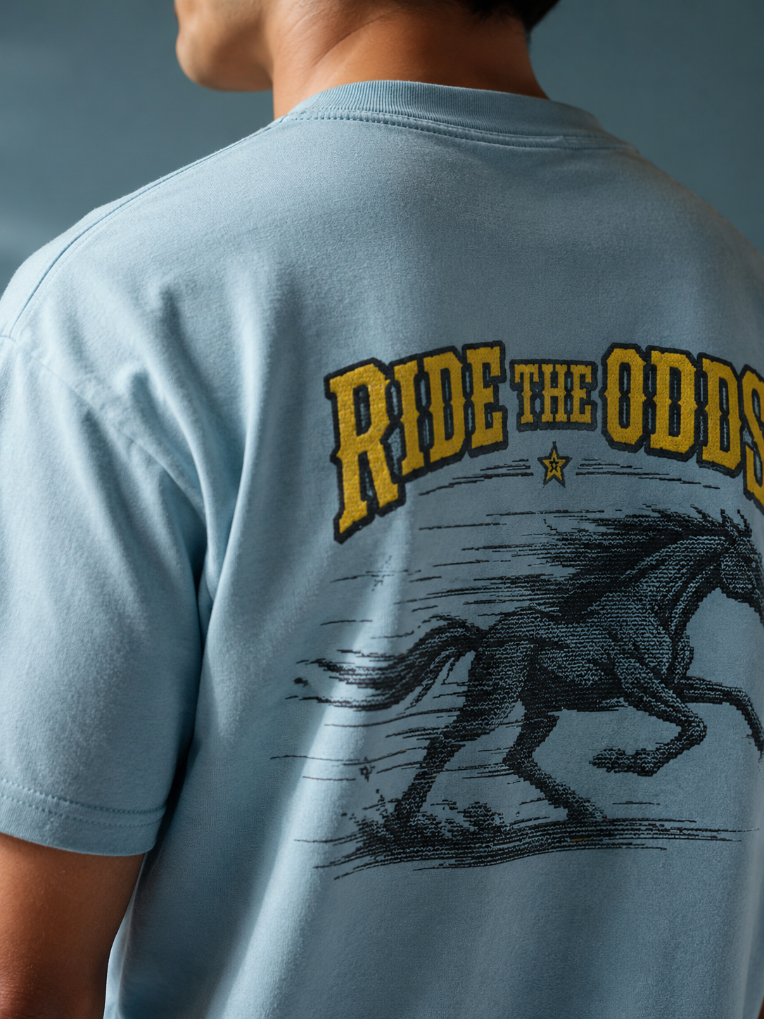 Close-up of the Ruxel Ride the Odds back print on light blue garment-dyed cotton - galloping black horse, star and gold RIDE THE ODDS western lettering