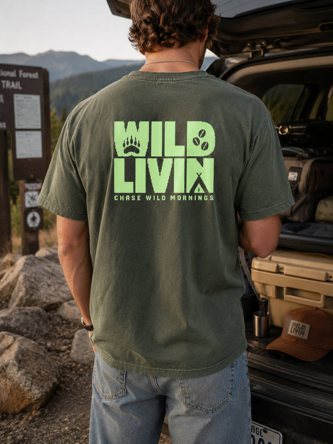 Man wearing the Ruxel Wild Livin olive graphic tee outdoors at a trailhead, back print shown