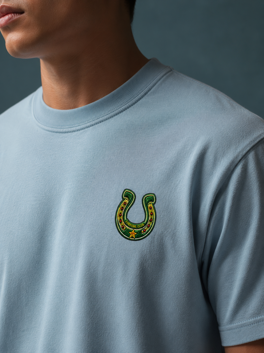 Close-up of the green Fortune Club horseshoe crest with gold star on the Ruxel Ride the Odds light blue garment-dyed tee