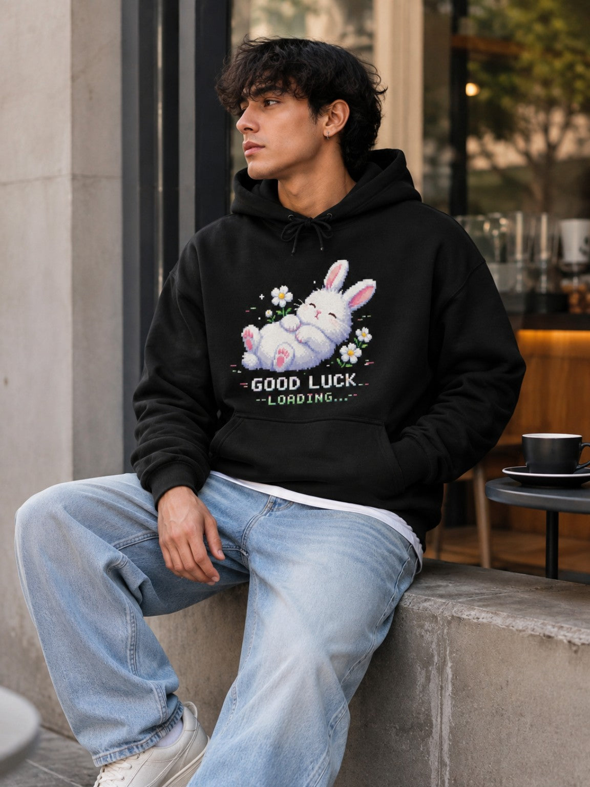 Male model wearing the Ruxel Good Luck Loading Void Black pixel bunny hoodie while seated outside a cafe