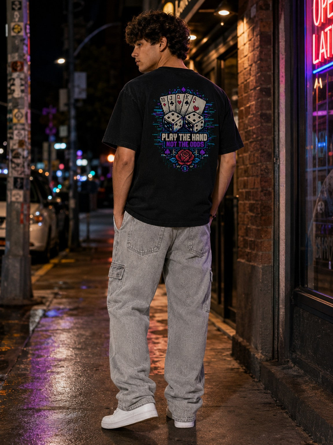 Back lifestyle view of the Ruxel Luck.exe Vintage Black pixel-art tee on a neon-lit city street, showing the full Play the Hand Not the Odds graphic