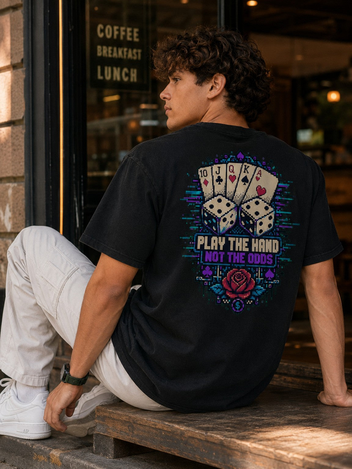 Seated lifestyle view of the Ruxel Luck.exe Play the Hand tee showing the oversized drape and full pixel-art back graphic